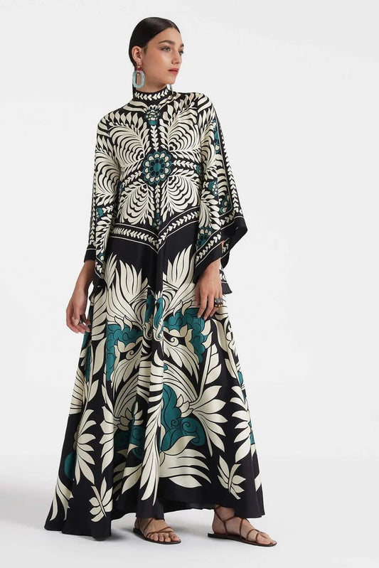 Multi - A woman wearing a high-neck maxi dress featuring a bold black, cream, and teal botanical medallion print, designed with dramatic cape sleeves and a fluid, floor-length skirt.