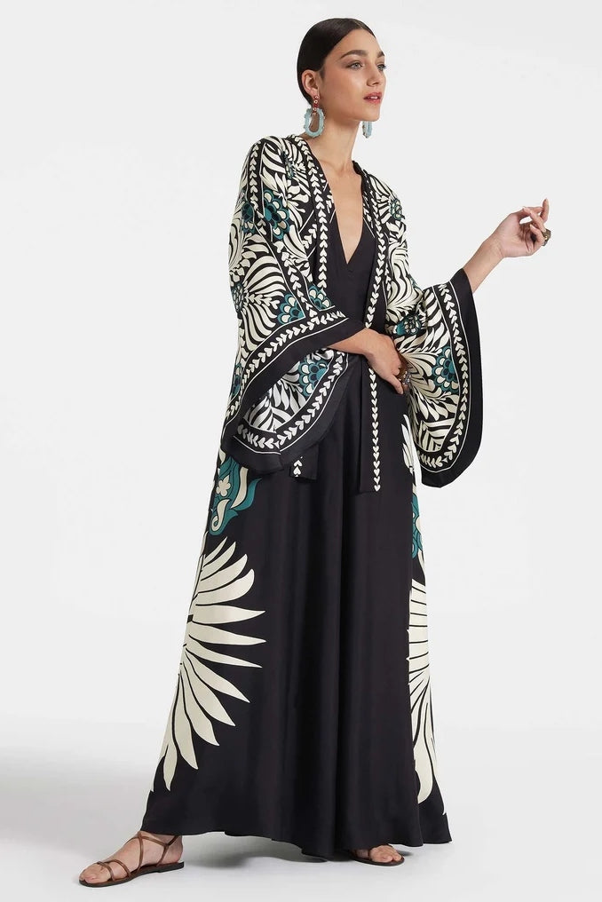 Multi - A woman wearing a high-neck maxi dress featuring a bold black, cream, and teal botanical medallion print, designed with dramatic cape sleeves and a fluid, floor-length skirt.