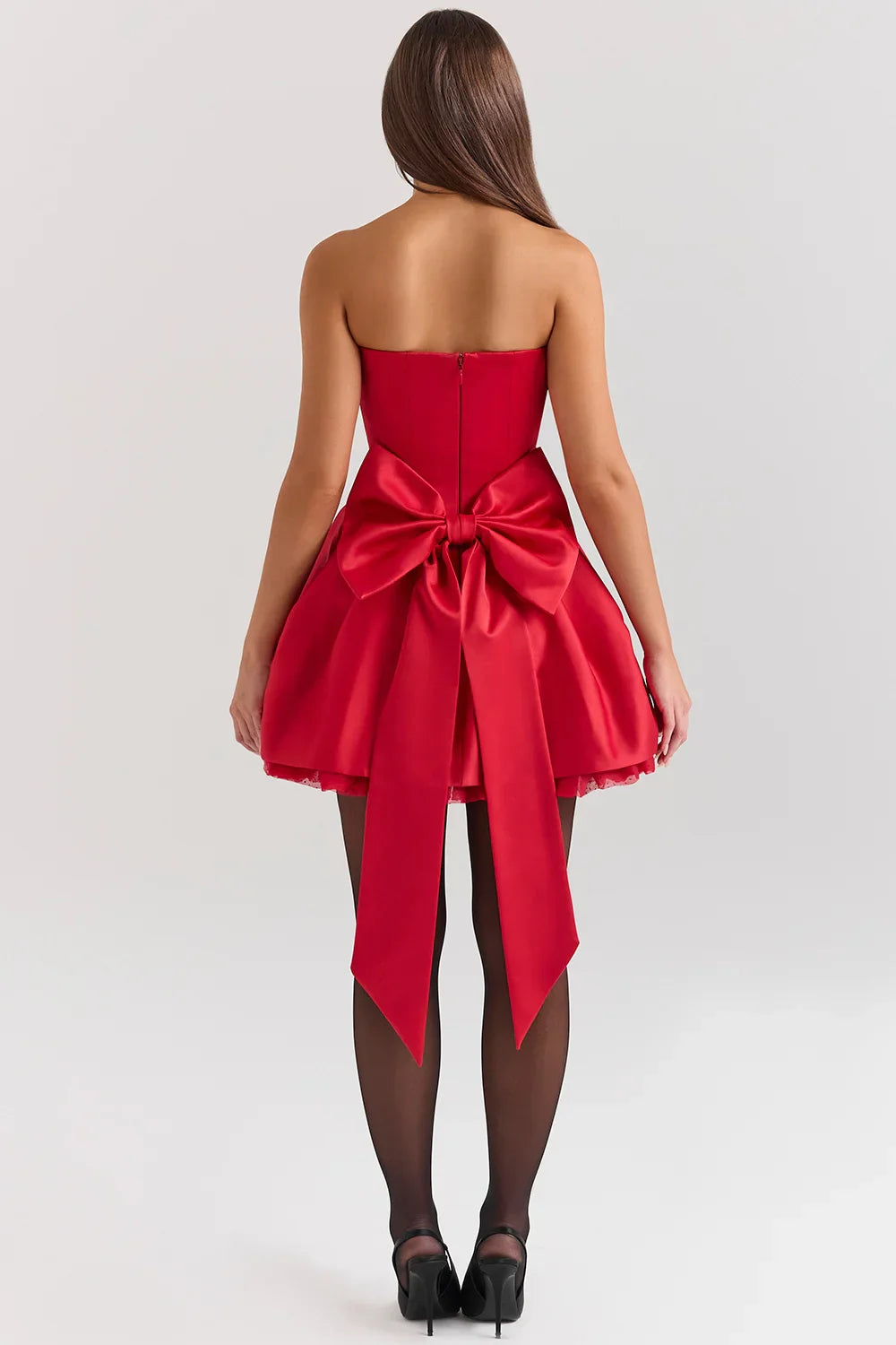 A woman models a vibrant red, strapless mini dress against a white background. The dress has a fitted corset bodice with a sweetheart neckline and a voluminous, pleated A-line skirt with a satin finish.