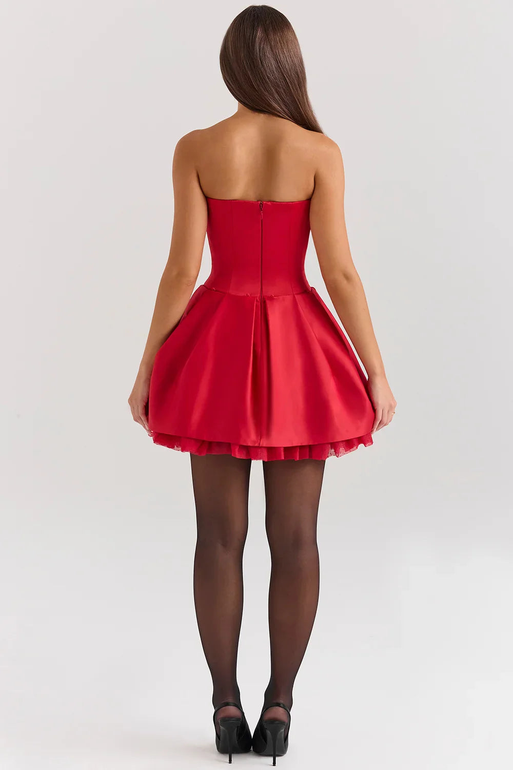 A woman models a vibrant red, strapless mini dress against a white background. The dress has a fitted corset bodice with a sweetheart neckline and a voluminous, pleated A-line skirt with a satin finish.