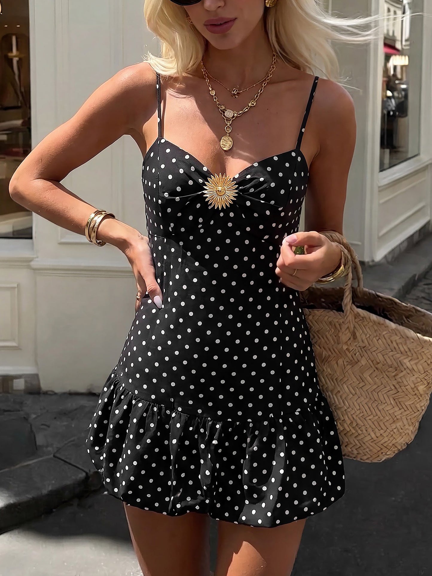 Black - Woman wearing a polka dot mini sundress with spaghetti straps, a ruched bust featuring a gold sunburst detail, and a ruffle hem, holding an ice cream cone outdoors.