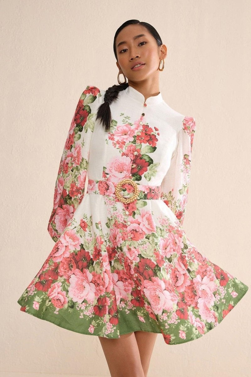 Multi - A woman with blonde hair in a sleek bun wears a long-sleeved, high-neck mini dress with a vibrant pink and red floral print, a matching belt, and a flared skirt, posing on a city street.