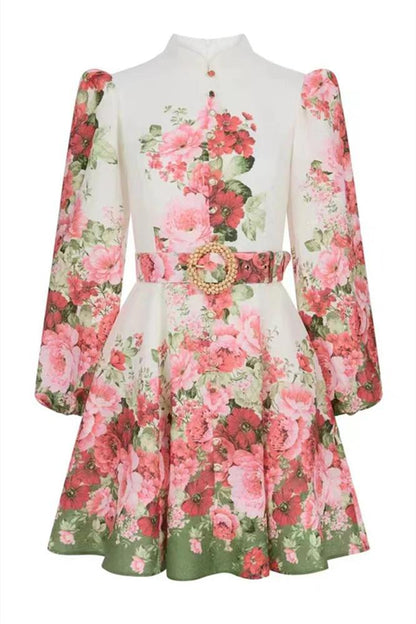 Multi - A woman with blonde hair in a sleek bun wears a long-sleeved, high-neck mini dress with a vibrant pink and red floral print, a matching belt, and a flared skirt, posing on a city street.