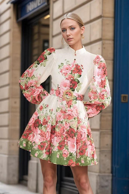 A woman with blonde hair in a sleek bun poses on a city street wearing a long-sleeve, mock-neck mini dress. The dress is off-white with a bold pink and red floral print, featuring voluminous puff sleeves, a matching belt with a large gold-tone buckle, and a flared A-line skirt.