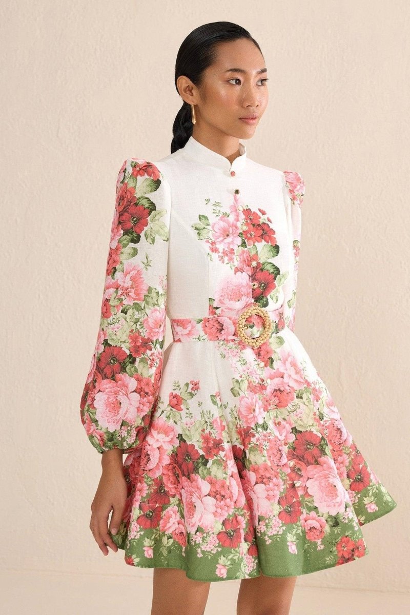 A woman with blonde hair in a sleek bun poses on a city street wearing a long-sleeve, mock-neck mini dress. The dress is off-white with a bold pink and red floral print, featuring voluminous puff sleeves, a matching belt with a large gold-tone buckle, and a flared A-line skirt.
