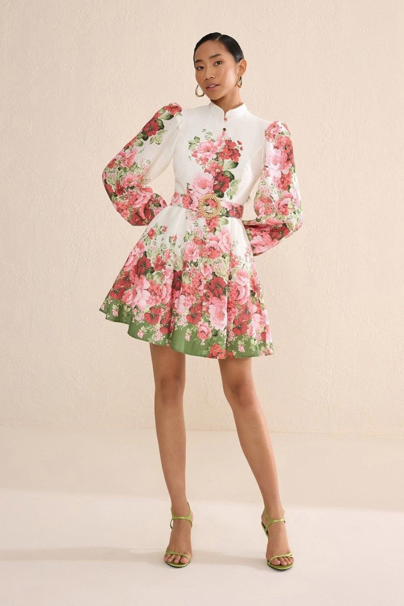 A woman with blonde hair in a sleek bun poses on a city street wearing a long-sleeve, mock-neck mini dress. The dress is off-white with a bold pink and red floral print, featuring voluminous puff sleeves, a matching belt with a large gold-tone buckle, and a flared A-line skirt.