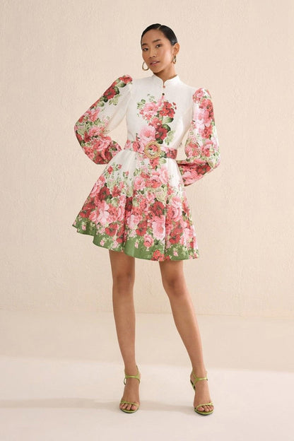Multi - A woman with blonde hair in a sleek bun wears a long-sleeved, high-neck mini dress with a vibrant pink and red floral print, a matching belt, and a flared skirt, posing on a city street.