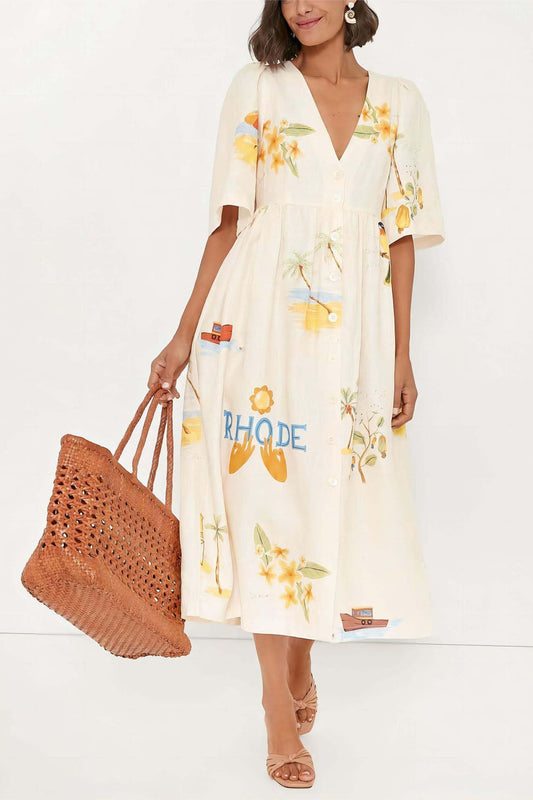 A woman wears a cream-colored, v-neck midi dress with elbow-length sleeves and a tropical vacation print. She holds a large brown woven tote bag and stands against a plain white background.