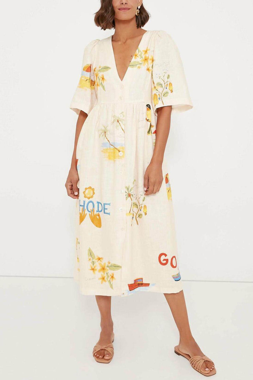 A woman wears a cream-colored, v-neck midi dress with elbow-length sleeves and a tropical vacation print. She holds a large brown woven tote bag and stands against a plain white background.