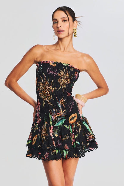 A woman models a strapless smocked mini dress featuring a vibrant tropical print on a black background. The dress has a form-fitting bodice and a short, tiered skirt with a scalloped hem.