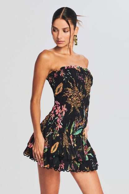 A woman models a strapless smocked mini dress featuring a vibrant tropical print on a black background. The dress has a form-fitting bodice and a short, tiered skirt with a scalloped hem.