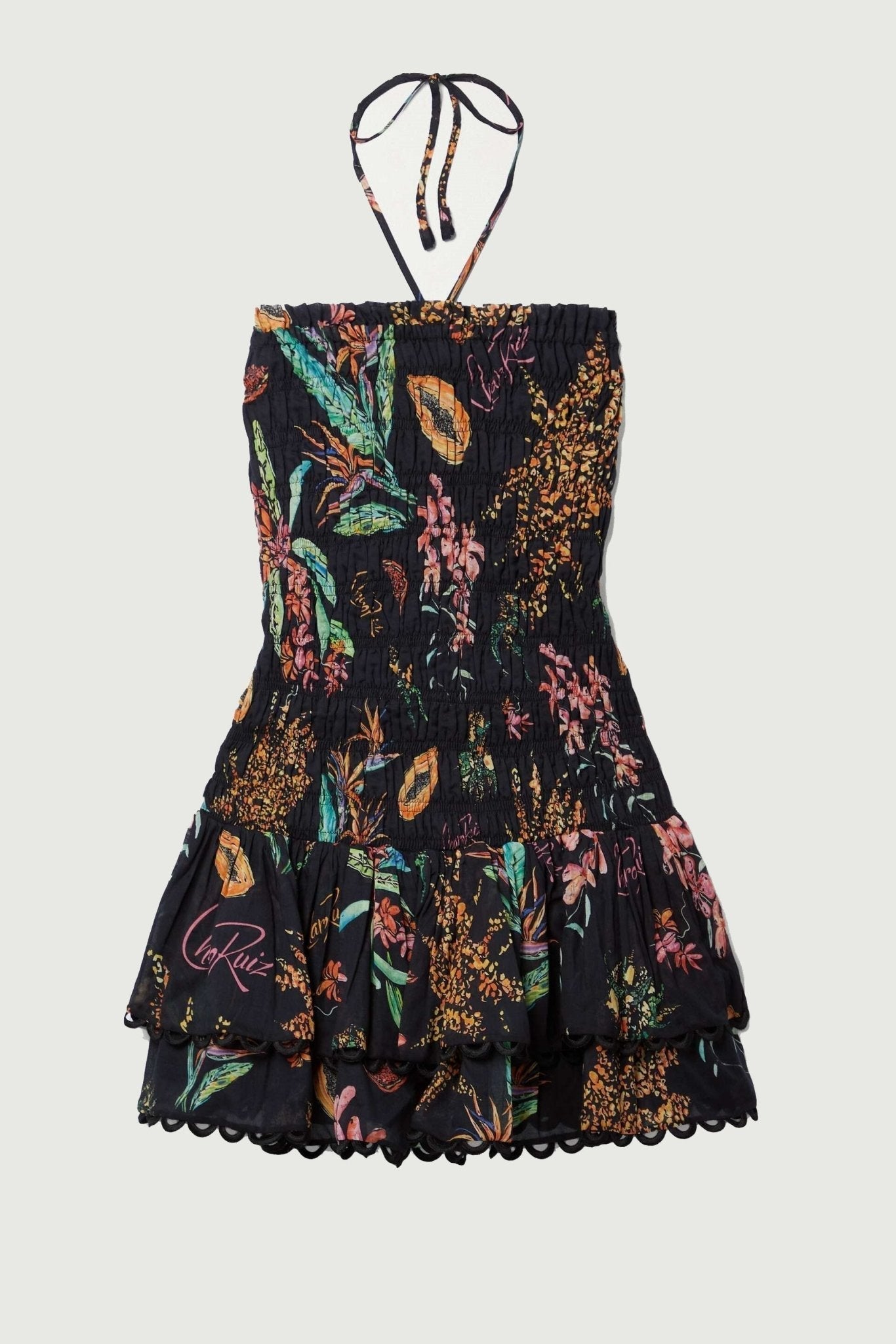 A woman models a strapless smocked mini dress featuring a vibrant tropical print on a black background. The dress has a form-fitting bodice and a short, tiered skirt with a scalloped hem.