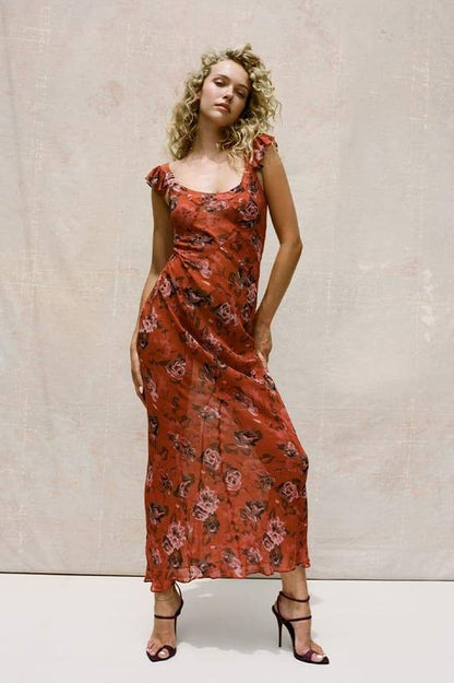 Red - A woman wearing a rust-colored, rose-printed chiffon maxi dress with ruffle cap sleeves and a scoop neckline, posing against a neutral textured backdrop.
