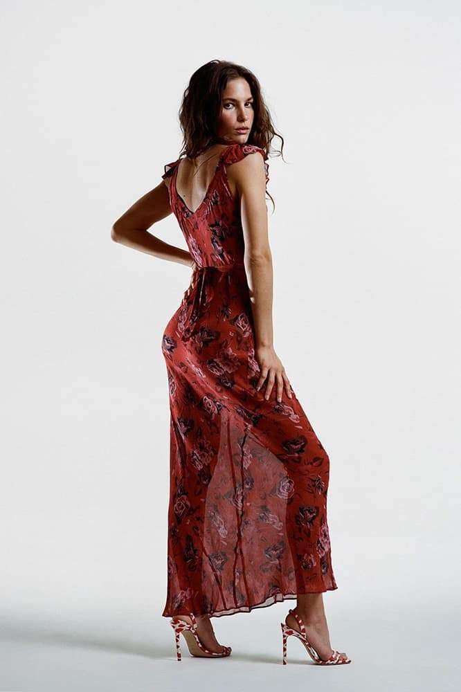 Red - A woman wearing a rust-colored, rose-printed chiffon maxi dress with ruffle cap sleeves and a scoop neckline, posing against a neutral textured backdrop.