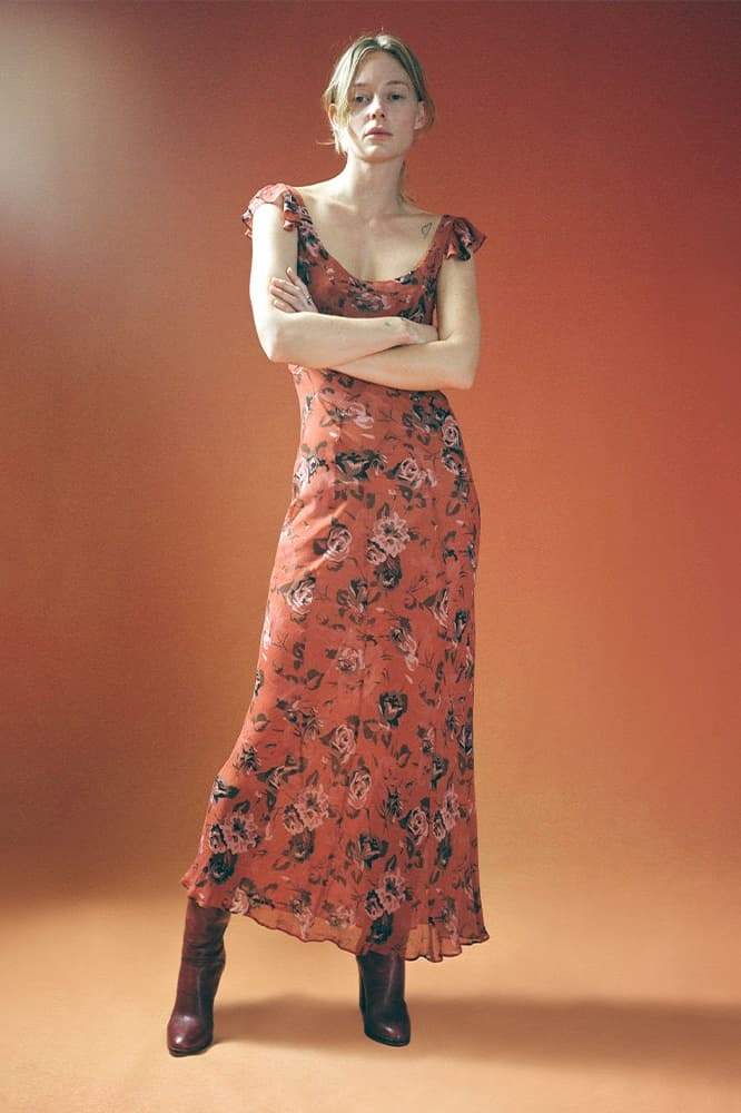 Red - A woman wearing a rust-colored, rose-printed chiffon maxi dress with ruffle cap sleeves and a scoop neckline, posing against a neutral textured backdrop.