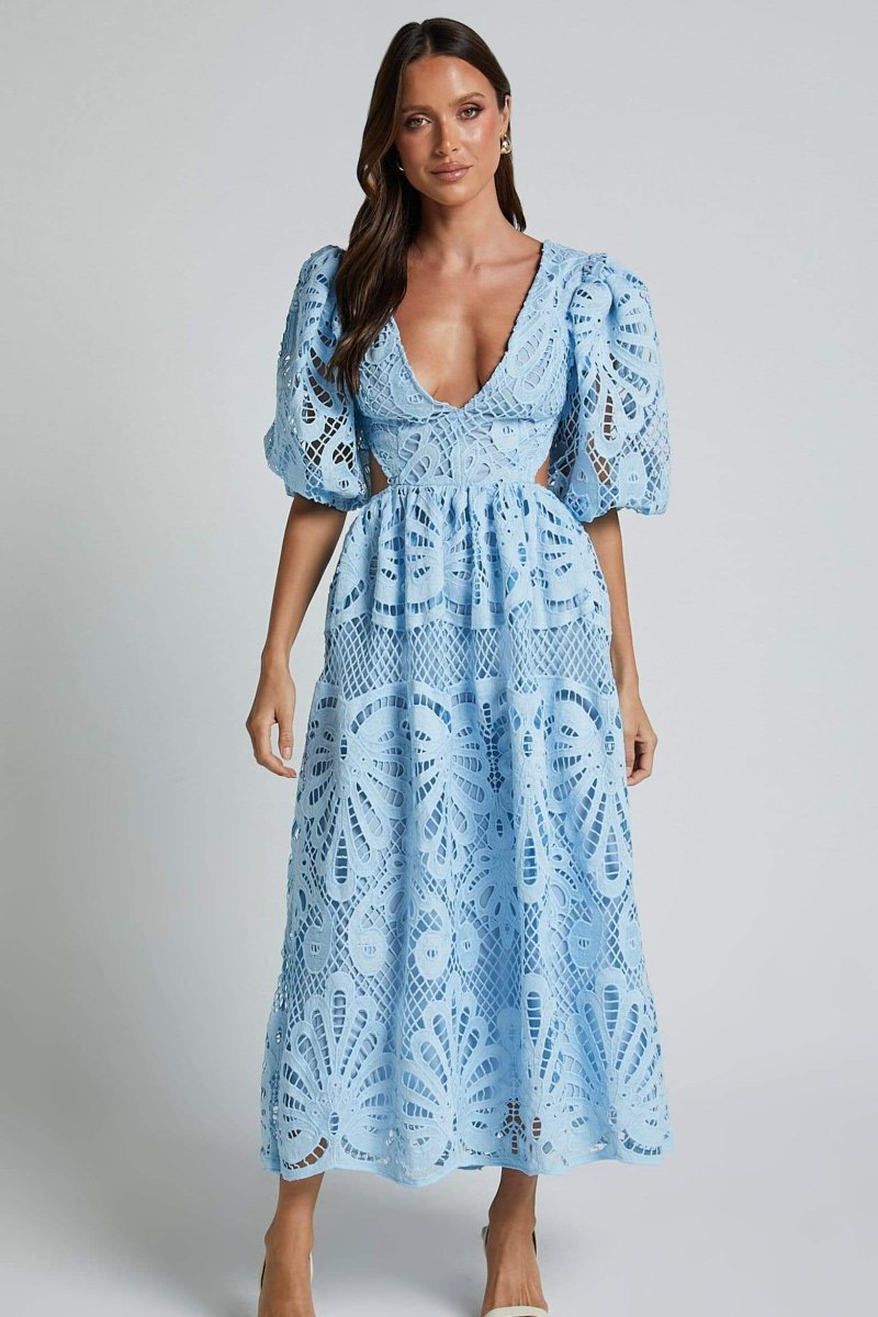 A woman with long brown hair models a yellow lace midi dress with a plunging V-neck, short puff sleeves, and side waist cutouts. She is holding a small beige handbag and wearing strappy heels, standing against a plain light gray background.
