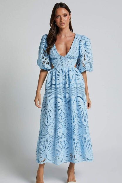 A woman with long brown hair models a yellow lace midi dress with a plunging V-neck, short puff sleeves, and side waist cutouts. She is holding a small beige handbag and wearing strappy heels, standing against a plain light gray background.