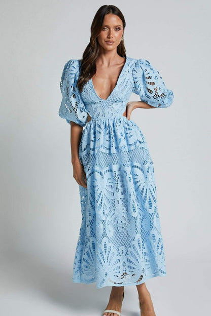 A woman with long brown hair models a yellow lace midi dress with a plunging V-neck, short puff sleeves, and side waist cutouts. She is holding a small beige handbag and wearing strappy heels, standing against a plain light gray background.