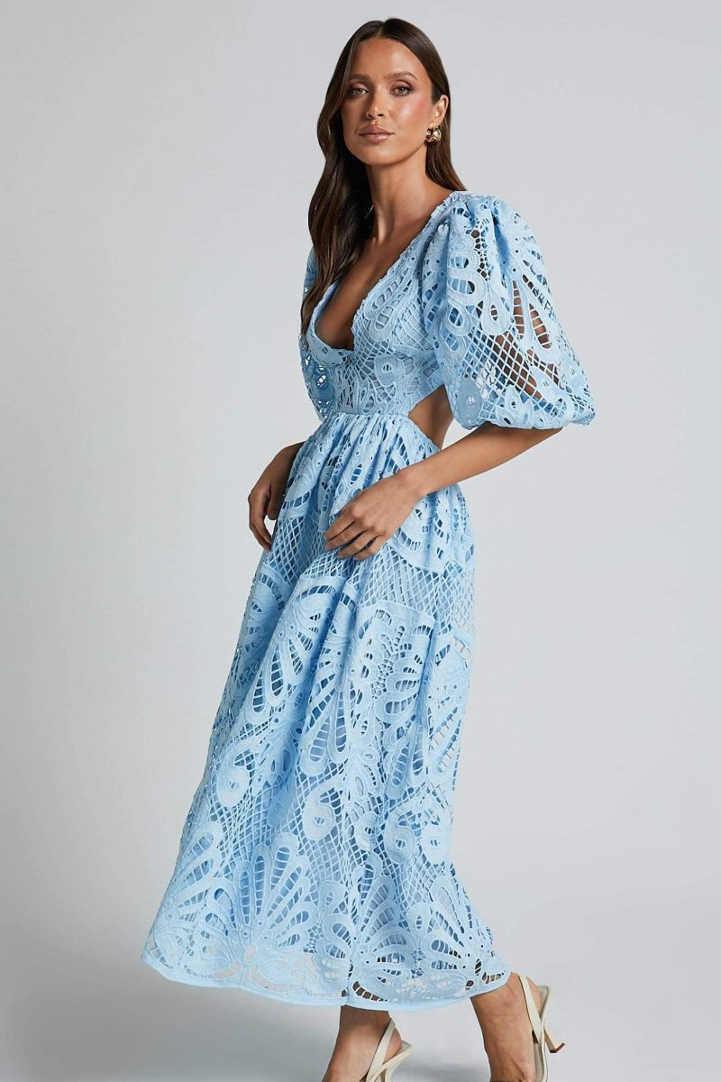 A woman with long brown hair models a yellow lace midi dress with a plunging V-neck, short puff sleeves, and side waist cutouts. She is holding a small beige handbag and wearing strappy heels, standing against a plain light gray background.
