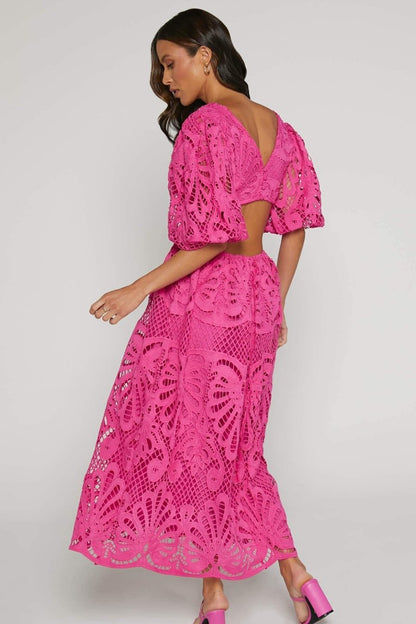 A woman with long brown hair models a yellow lace midi dress with a plunging V-neck, short puff sleeves, and side waist cutouts. She is holding a small beige handbag and wearing strappy heels, standing against a plain light gray background.