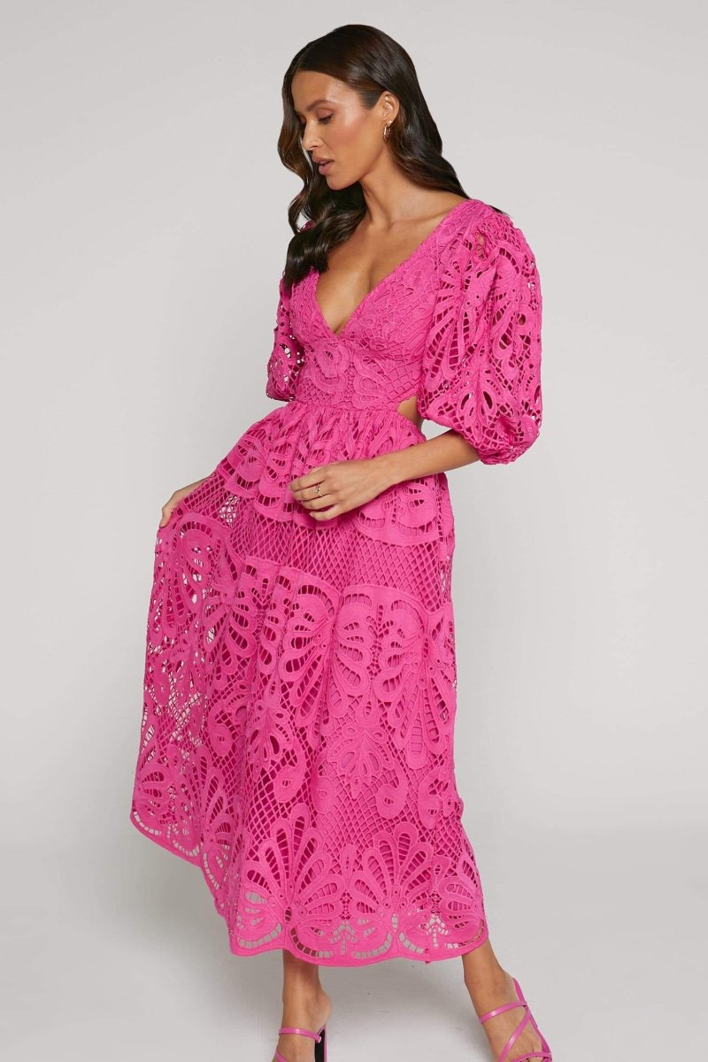 A woman with long brown hair models a yellow lace midi dress with a plunging V-neck, short puff sleeves, and side waist cutouts. She is holding a small beige handbag and wearing strappy heels, standing against a plain light gray background.