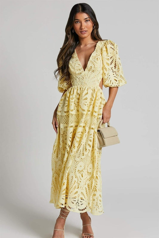 A woman with long brown hair models a yellow lace midi dress with a plunging V-neck, short puff sleeves, and side waist cutouts. She is holding a small beige handbag and wearing strappy heels, standing against a plain light gray background.