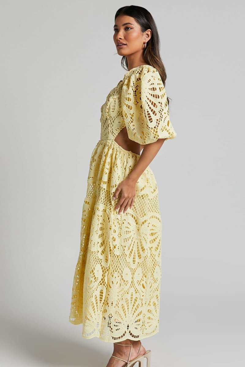 A woman with long brown hair models a yellow lace midi dress with a plunging V-neck, short puff sleeves, and side waist cutouts. She is holding a small beige handbag and wearing strappy heels, standing against a plain light gray background.