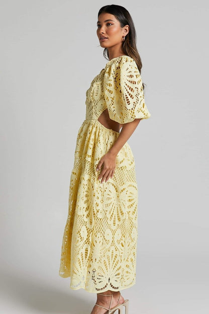 A woman with long brown hair models a yellow lace midi dress with a plunging V-neck, short puff sleeves, and side waist cutouts. She is holding a small beige handbag and wearing strappy heels, standing against a plain light gray background.