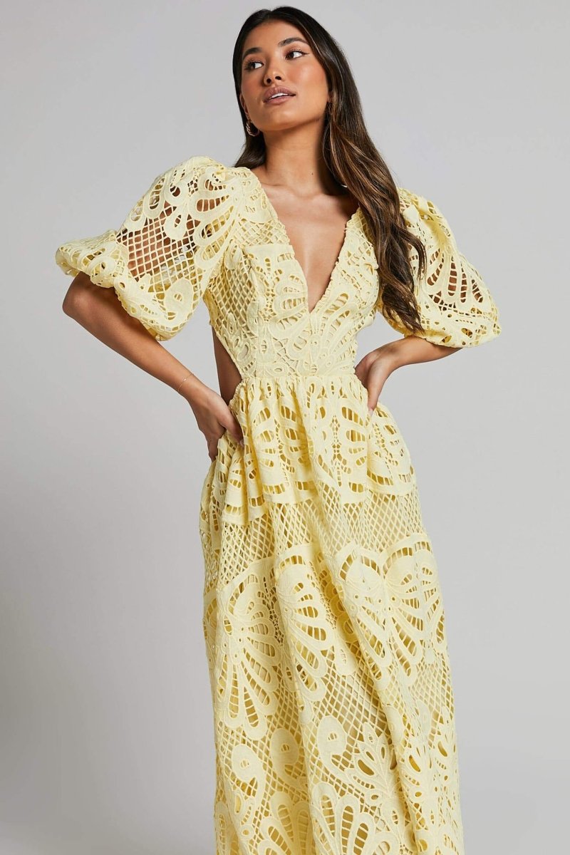 A woman with long brown hair models a yellow lace midi dress with a plunging V-neck, short puff sleeves, and side waist cutouts. She is holding a small beige handbag and wearing strappy heels, standing against a plain light gray background.