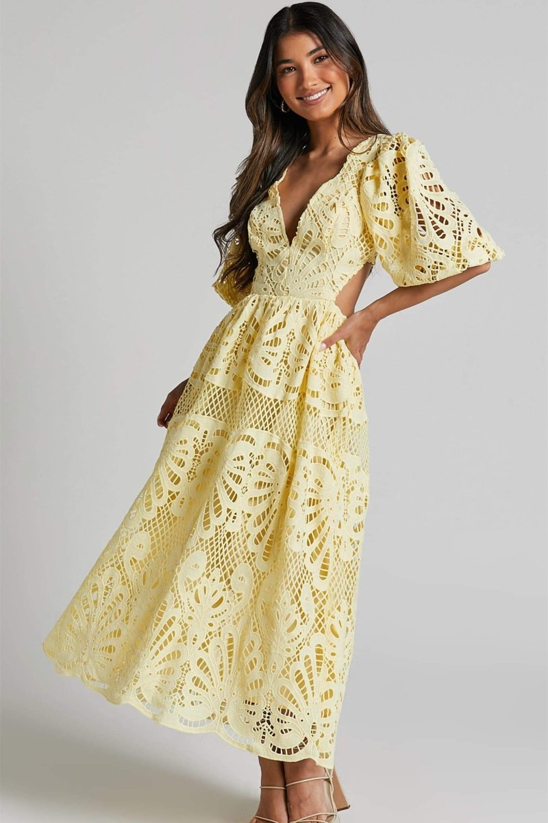 A woman with long brown hair models a yellow lace midi dress with a plunging V-neck, short puff sleeves, and side waist cutouts. She is holding a small beige handbag and wearing strappy heels, standing against a plain light gray background.