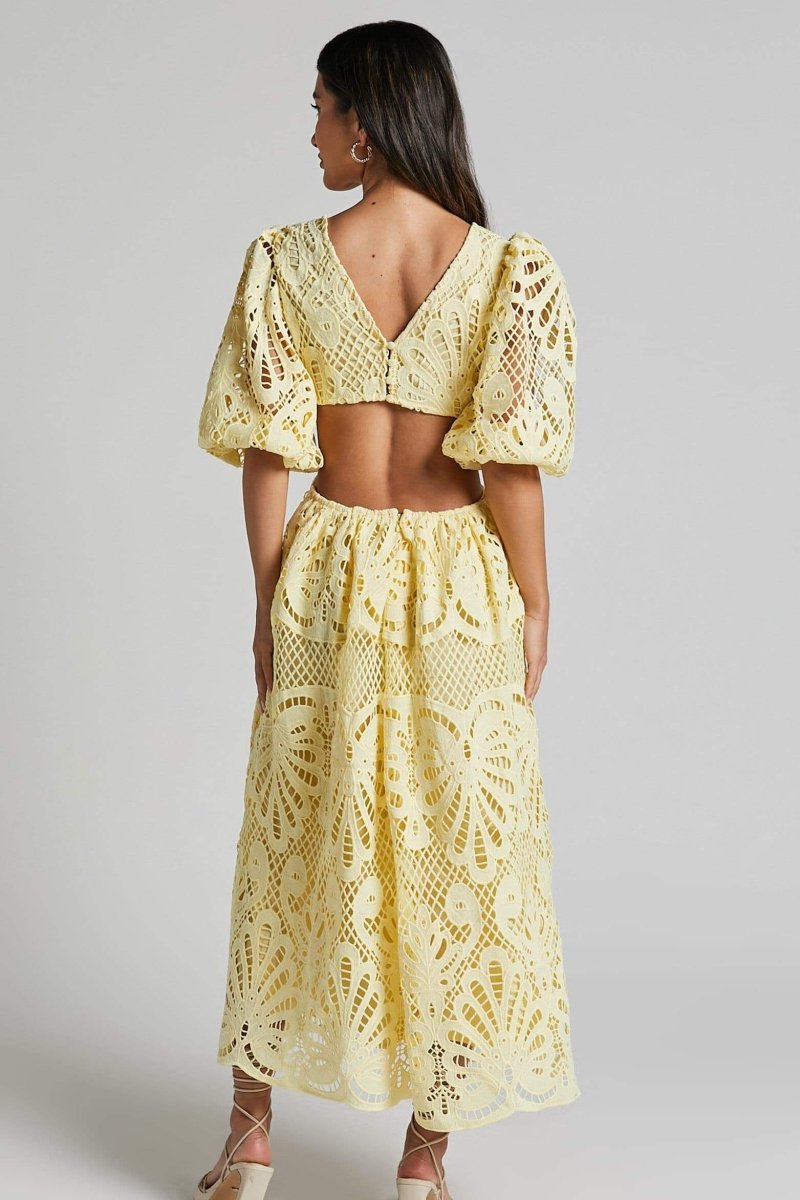 A woman with long brown hair models a yellow lace midi dress with a plunging V-neck, short puff sleeves, and side waist cutouts. She is holding a small beige handbag and wearing strappy heels, standing against a plain light gray background.