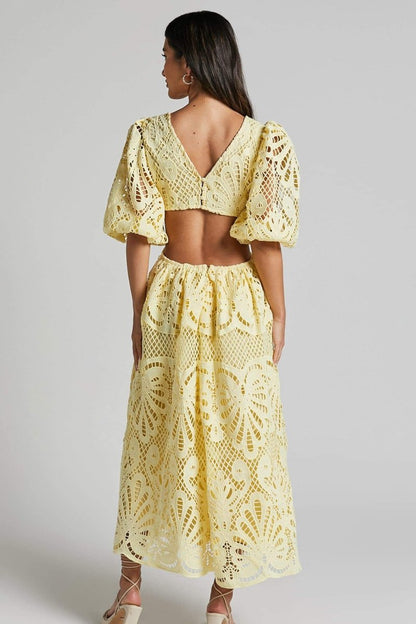 A woman with long brown hair models a yellow lace midi dress with a plunging V-neck, short puff sleeves, and side waist cutouts. She is holding a small beige handbag and wearing strappy heels, standing against a plain light gray background.
