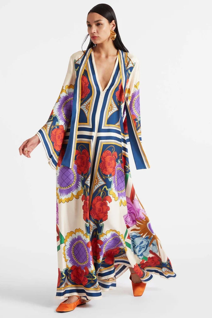A model wears a floor-length satin maxi dress featuring a bold, colorful floral and geometric scarf print in cream, blue, red, and purple, designed with a deep V-neckline, long scarf collar, and wide flowing sleeves.
