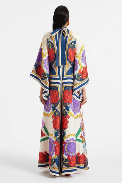 A model wears a floor-length satin maxi dress featuring a bold, colorful floral and geometric scarf print in cream, blue, red, and purple, designed with a deep V-neckline, long scarf collar, and wide flowing sleeves.