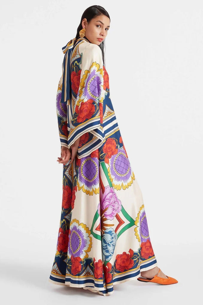 Multi - A model wears a floor-length satin maxi dress featuring a bold, colorful floral and geometric scarf print in cream, blue, red, and purple, designed with a deep V-neckline, long scarf collar, and wide flowing sleeves.