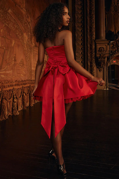 A woman models a vibrant red, strapless mini dress against a white background. The dress has a fitted corset bodice with a sweetheart neckline and a voluminous, pleated A-line skirt with a satin finish.
