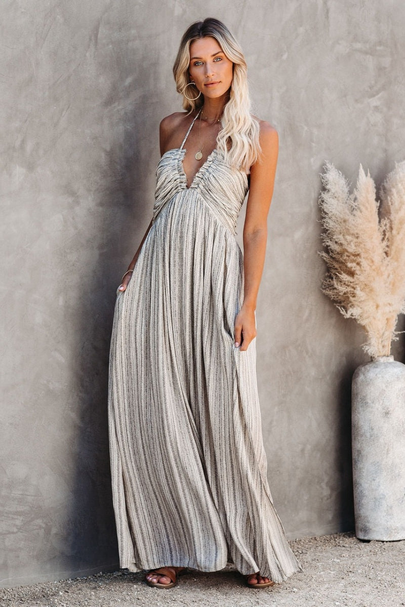 Grey - A woman wearing a sleeveless halter maxi dress with a textured micro-stripe pattern, featuring a ruched bust and side slits, posed against a rustic concrete wall.