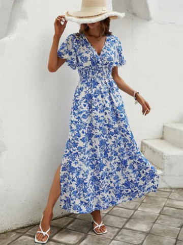 XL - A woman wearing a blue and white toile floral print chiffon maxi dress with flutter sleeves, a smocked waist, and a side slit, paired with a wide-brim straw hat and white strappy sandals.