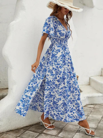 XL - A woman wearing a blue and white toile floral print chiffon maxi dress with flutter sleeves, a smocked waist, and a side slit, paired with a wide-brim straw hat and white strappy sandals.