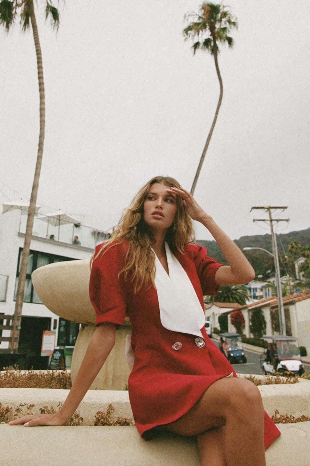 A woman with long blonde hair leans against a metal pole, wearing a structured red mini blazer dress with short puff sleeves, a wide white contrast lapel, and a flared A-line skirt.
