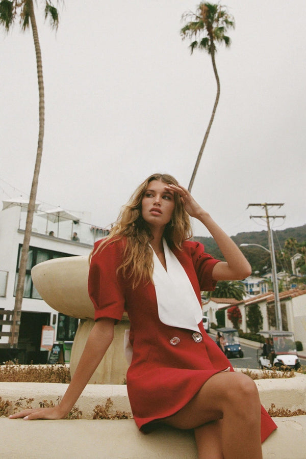 A woman with long blonde hair leans against a metal pole, wearing a structured red mini blazer dress with short puff sleeves, a wide white contrast lapel, and a flared A-line skirt.