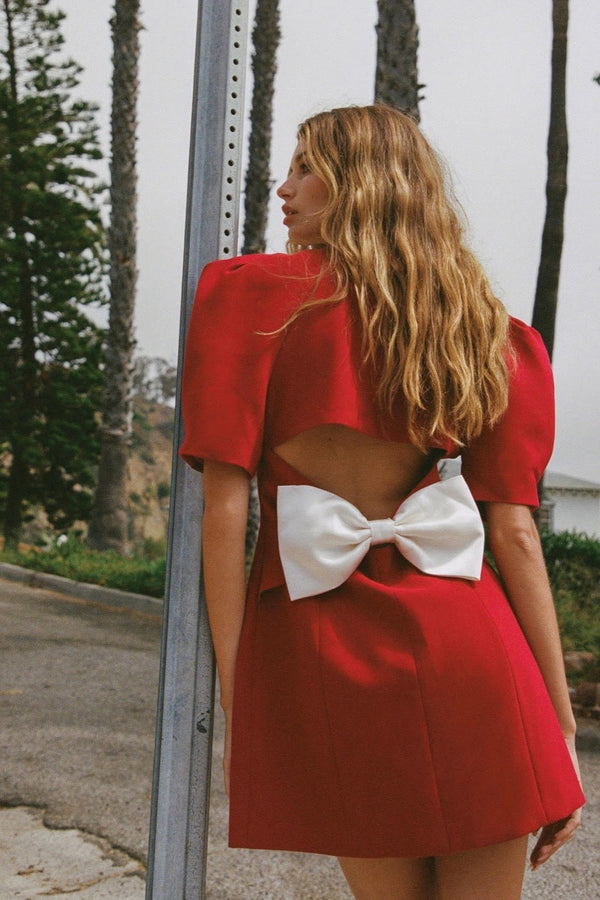 A woman with long blonde hair leans against a metal pole, wearing a structured red mini blazer dress with short puff sleeves, a wide white contrast lapel, and a flared A-line skirt.