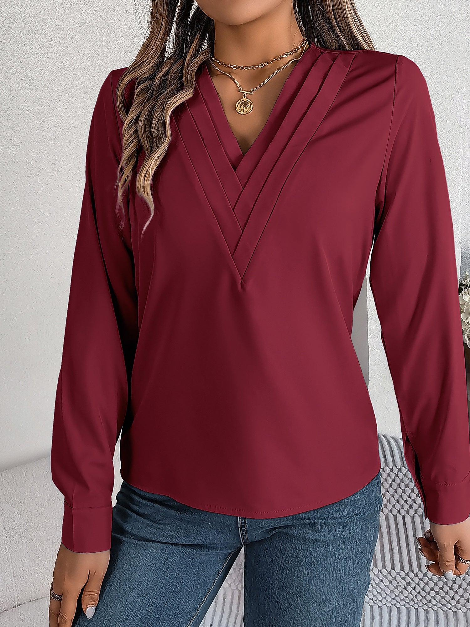 Burgundy - Women's long-sleeve blouse featuring a distinctive pleated V-neckline and buttoned cuffs, styled tucked into blue denim jeans.