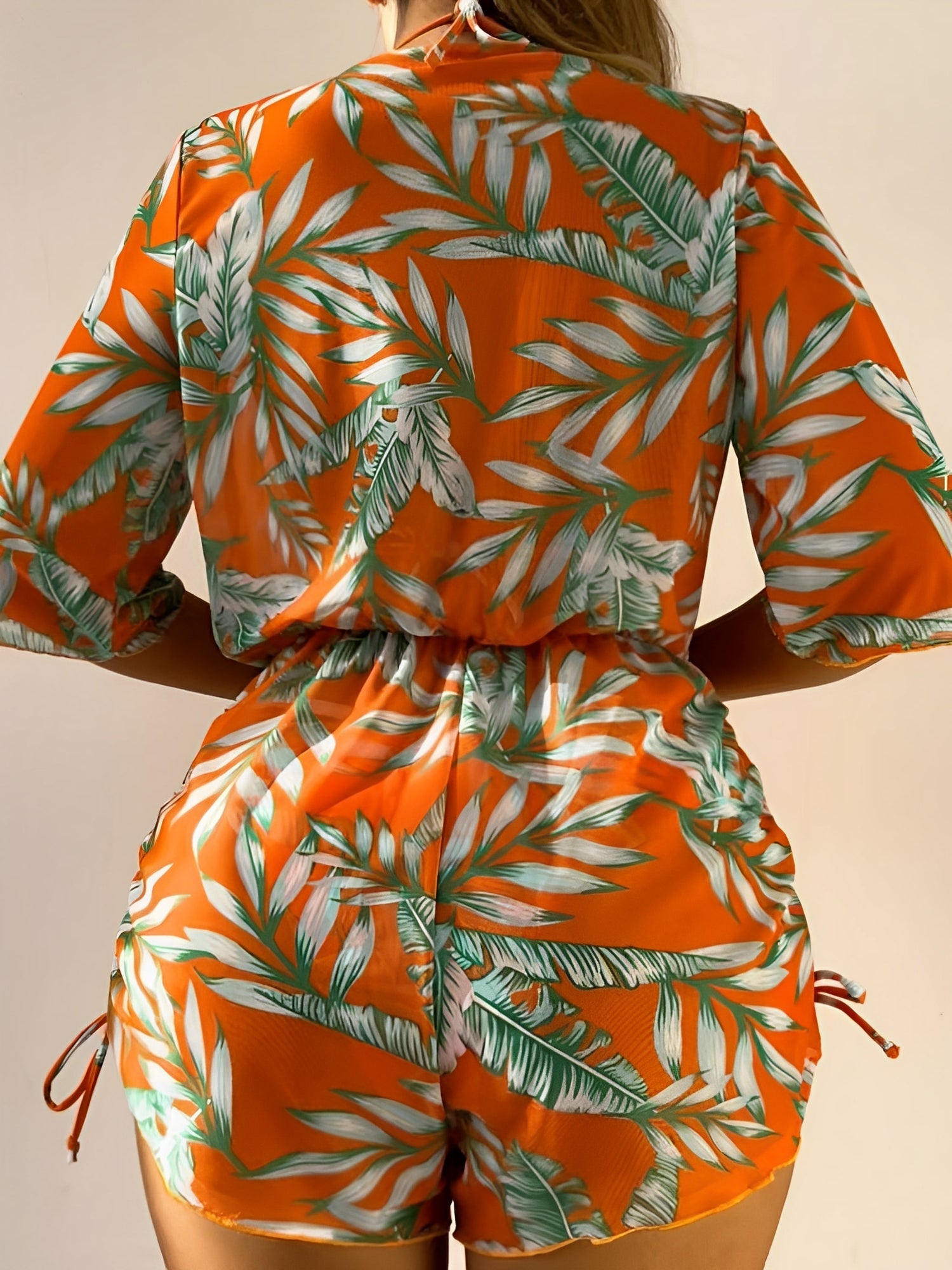 Orange - A woman models a sheer, short-sleeve mini cover-up with a vibrant tropical leaf print in shades of purple, blue, and pink, featuring a deep V-neck, drawstring waist, and ruched sides with ties.