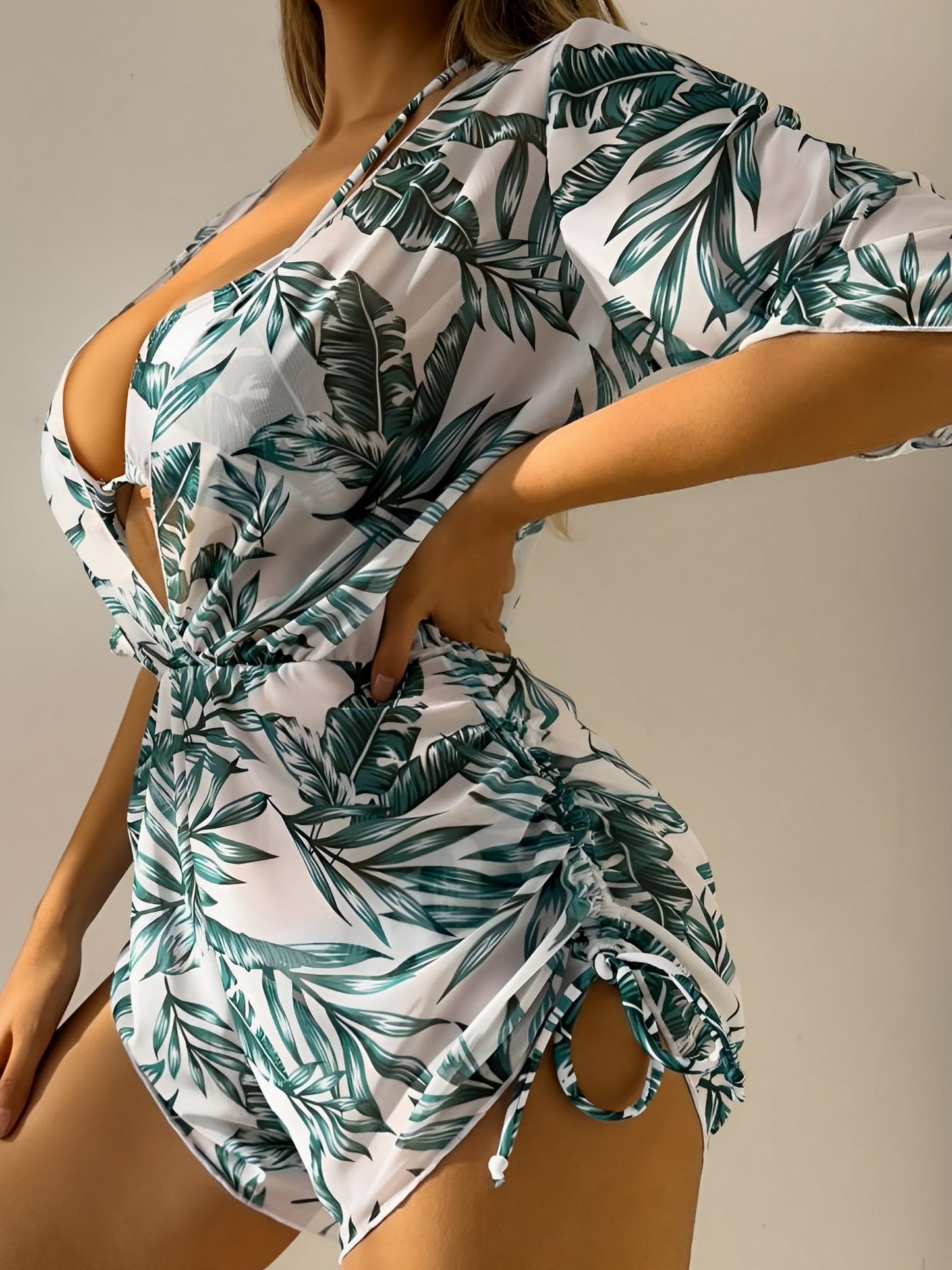Green - A woman models a sheer, short-sleeve mini cover-up with a vibrant tropical leaf print in shades of purple, blue, and pink, featuring a deep V-neck, drawstring waist, and ruched sides with ties.