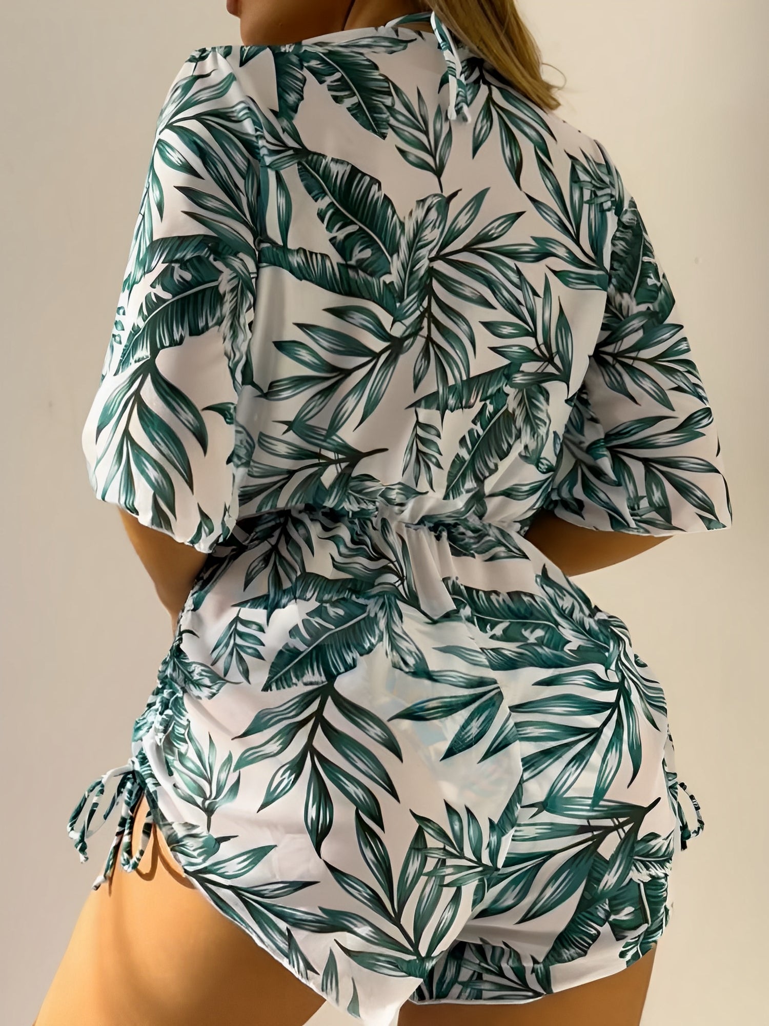 Green - A woman models a sheer, short-sleeve mini cover-up with a vibrant tropical leaf print in shades of purple, blue, and pink, featuring a deep V-neck, drawstring waist, and ruched sides with ties.
