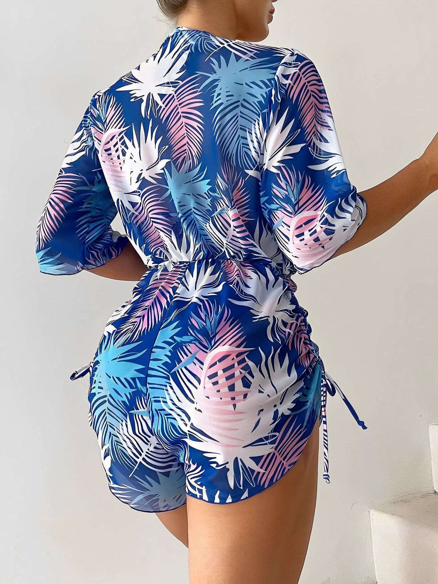 Royal Blue - A woman models a sheer, short-sleeve mini cover-up with a vibrant tropical leaf print in shades of purple, blue, and pink, featuring a deep V-neck, drawstring waist, and ruched sides with ties.