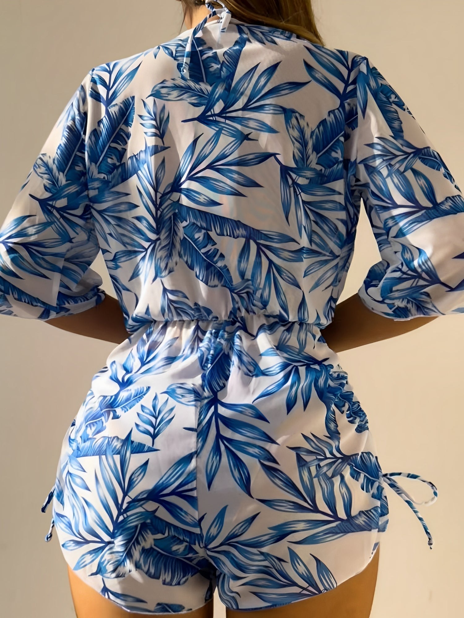 Blue - A woman models a sheer, short-sleeve mini cover-up with a vibrant tropical leaf print in shades of purple, blue, and pink, featuring a deep V-neck, drawstring waist, and ruched sides with ties.