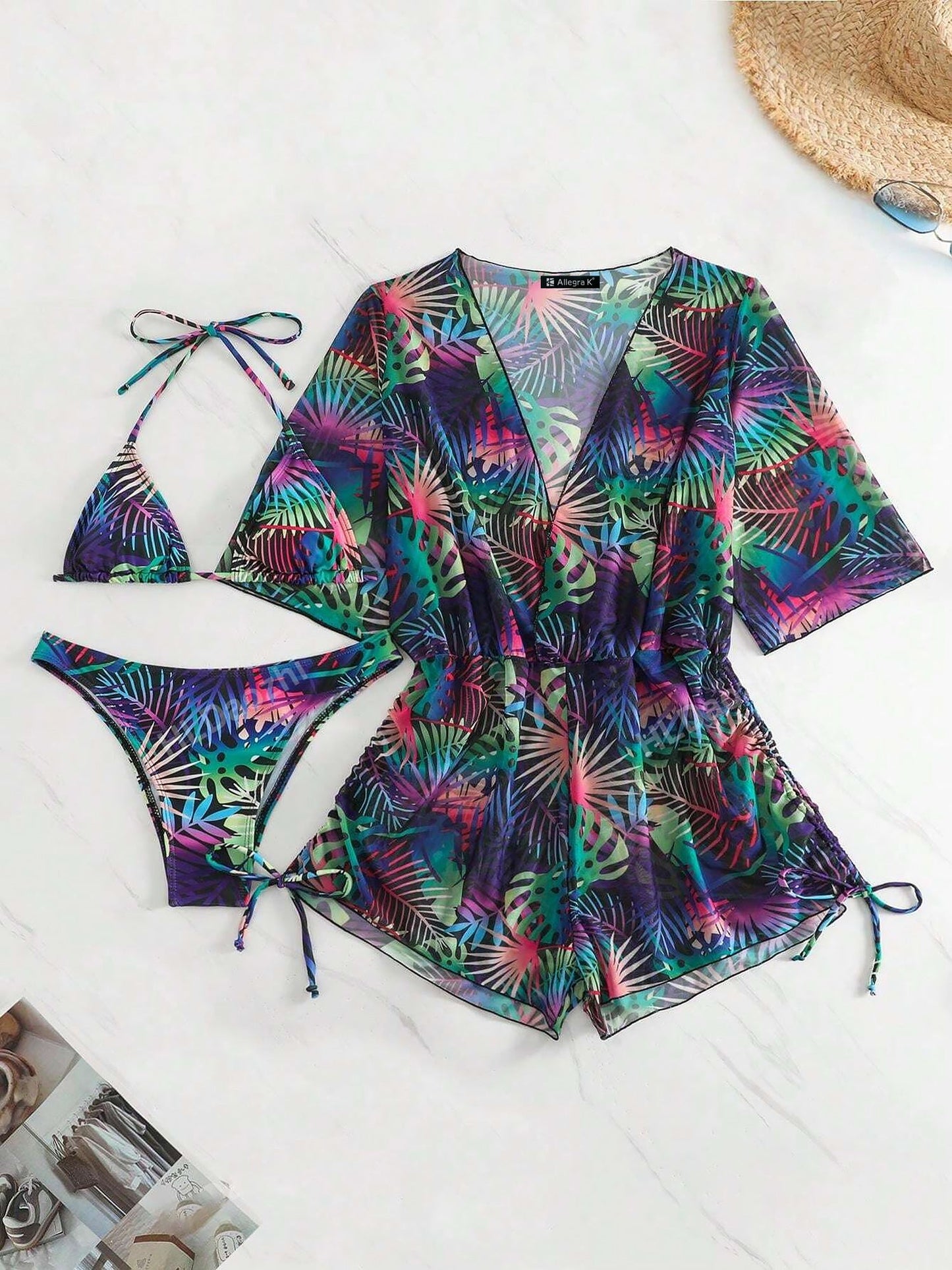 Purple - A woman models a sheer, short-sleeve mini cover-up with a vibrant tropical leaf print in shades of purple, blue, and pink, featuring a deep V-neck, drawstring waist, and ruched sides with ties.