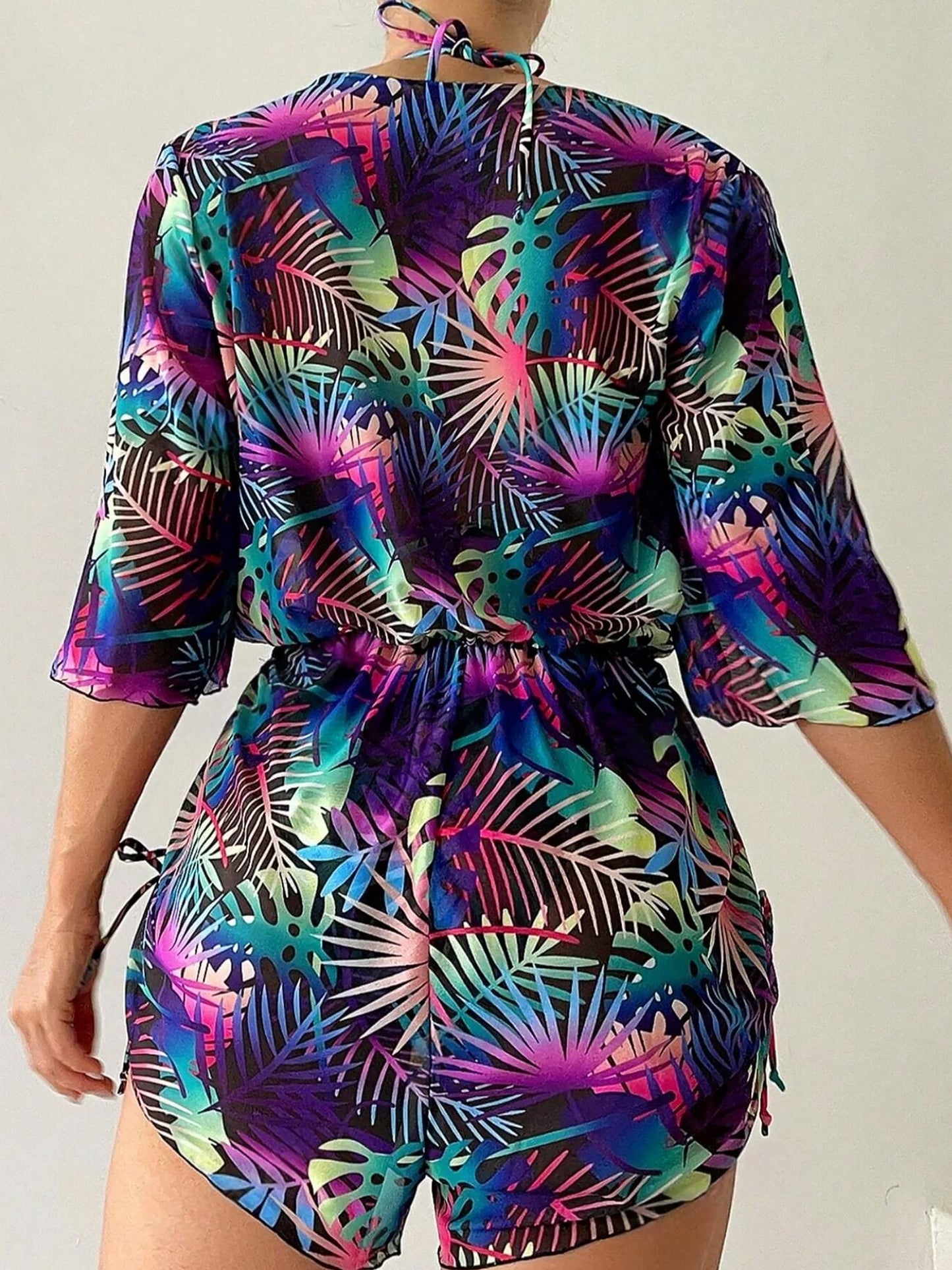Purple - A woman models a sheer, short-sleeve mini cover-up with a vibrant tropical leaf print in shades of purple, blue, and pink, featuring a deep V-neck, drawstring waist, and ruched sides with ties.